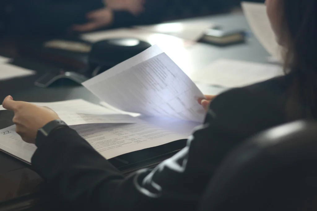 Close-up over-the-shoulder view of a person flipping through printed documents with a table schedule, in a dimly lit meeting room with other people in the background.