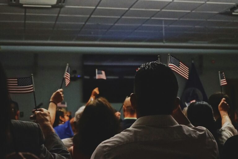 View from behind a seated crowd indoors, several people raising small American flags in the air, at a naturalization.