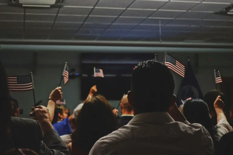 View from behind a seated crowd indoors, several people raising small American flags in the air, at a naturalization.
