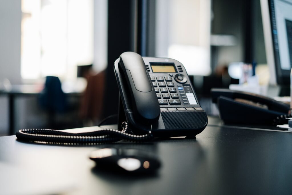 Close-up of a black desk phone with keypad and coiled handset cord on an office desk, with a blurred computer mouse in the foreground and an open workspace in the background.