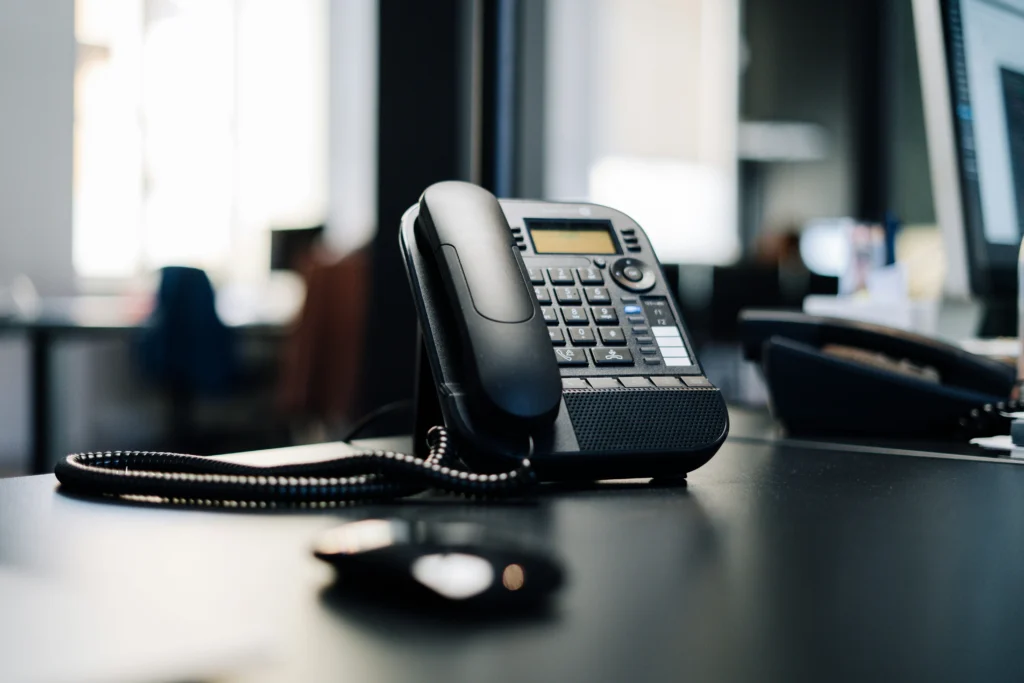 Close-up of a black desk phone with keypad and coiled handset cord on an office desk, with a blurred computer mouse in the foreground and an open workspace in the background.