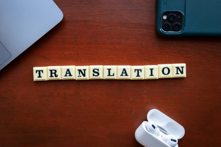 Overhead view of letter tiles spelling 'TRANSLATION' on a wooden desk, surrounded by a laptop, smartphone, and wireless earbuds in their open case.