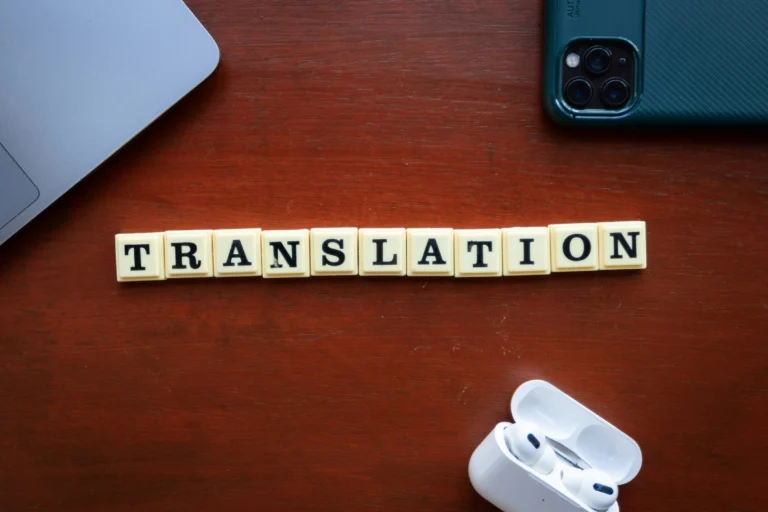 Overhead view of letter tiles spelling 'TRANSLATION' on a wooden desk, surrounded by a laptop, smartphone, and wireless earbuds in their open case.