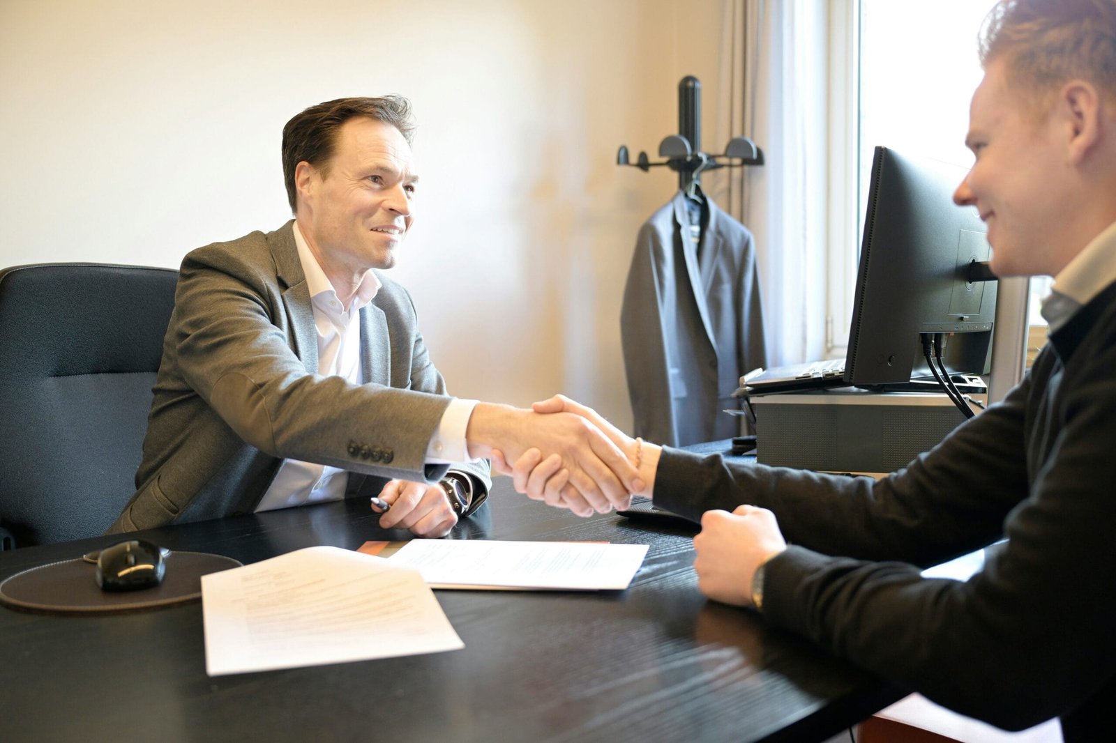 Two men in business attire shaking hands across a dark office desk, with documents and a computer monitor visible in the background.