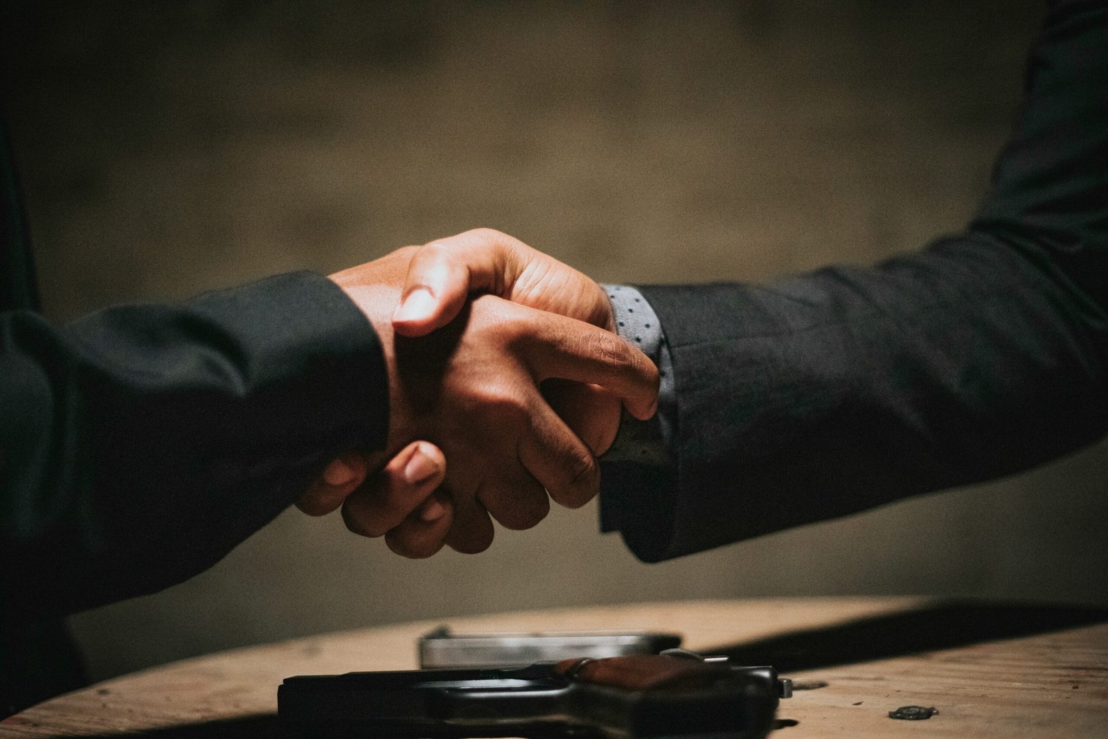 Close-up of two people in suits shaking hands above a wooden table with a handgun resting on it, suggesting a tense or dangerous deal.