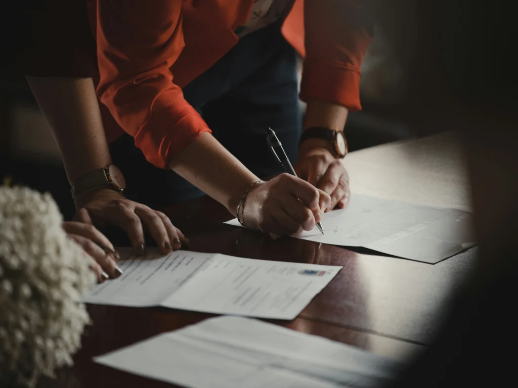 Close-up of a woman's hand signing a document on a wooden table, with additional papers and a dried floral arrangement in the foreground, lit by soft natural light.