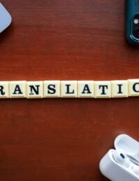 Overhead view of letter tiles spelling 'TRANSLATION' on a wooden desk, surrounded by a laptop, smartphone, and wireless earbuds in their open case.